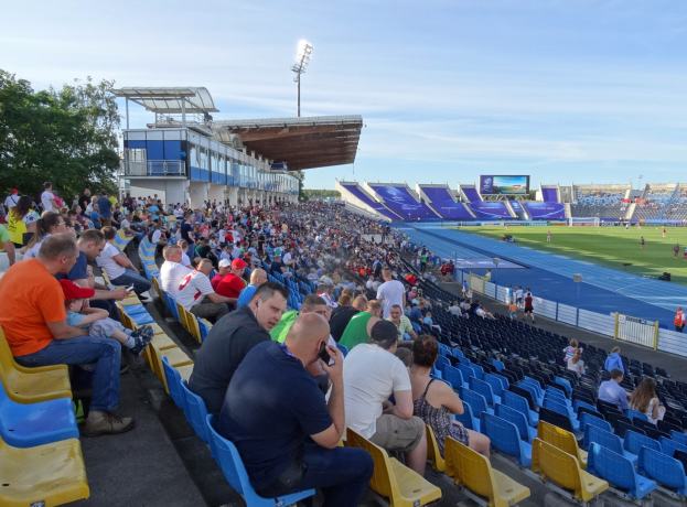 Eine große Menschenmenge in einem Stadion mit einem Zaun, Anzeigetafel, Dach mit Metallstangen, Bäumen und einem bewölkten Himmel, die ein Fußballspiel sehen.
