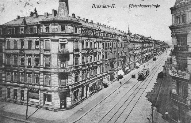 Ein altes Schwarz-Weiß-Foto einer Straßenkreuzung in Dresden, Deutschland, mit Fahrzeugen, Passanten, Gebäuden mit Fenstern und Schildern, Strommasten mit Kabeln und einem Himmel im Hintergrund, mit Text oben und unten auf dem Bild.