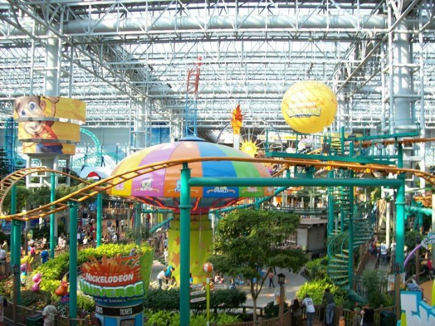 Ein großer Indoor-Wasserpark mit einer Achterbahn, umgeben von Menschen, Bäumen, Pflanzen und Deckenleuchten, die eine lebendige Atmosphäre schaffen.