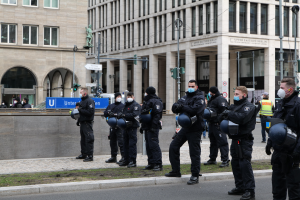 Eine Gruppe von Polizisten in schwarzen Uniformen und Masken steht vor einem Berliner Gebäude mit Glasfenstern, Säulen und grasbewachsenem Boden, einige halten Helme, während Laternen, Verkehrszeichen, Schilder mit Text, eine Statue und andere Menschen im Hintergrund zu sehen sind.