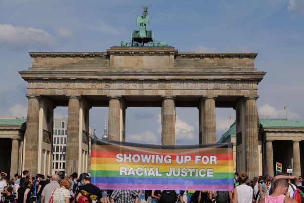 Gruppe von Menschen vor dem Brandenburger Tor in Berlin, Deutschland, mit einer "Racial Justice"-Schleife, die die Pfeiler und die Statue des Tors zeigt, sowie im Hintergrund Gebäude und bewölktem Himmel.