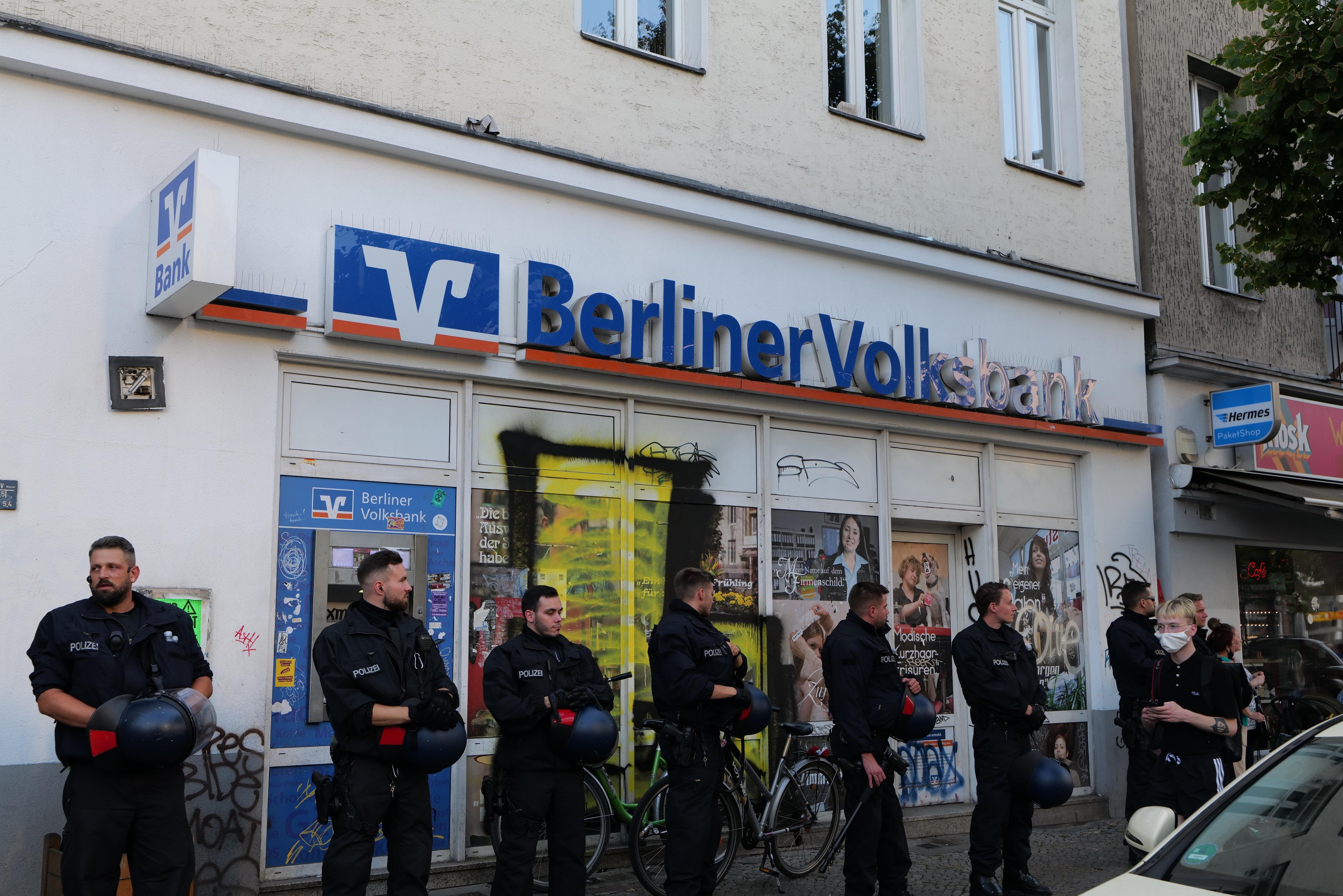 Gruppe von Polizeibeamten vor einer Berliner Volksbank-Filiale stehend, einige mit Helmen in den Händen, daneben ein Auto und ein Baum; im Hintergrund Gebäude mit Fenstern und Schildern.
