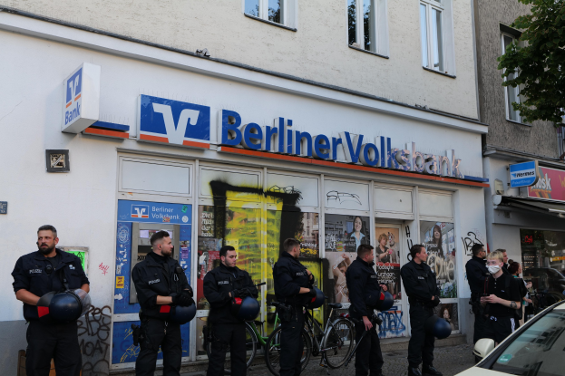 Gruppe von Polizeibeamten vor einer Berliner Volksbank-Filiale stehend, einige mit Helmen in den Händen, daneben ein Auto und ein Baum; im Hintergrund Gebäude mit Fenstern und Schildern.