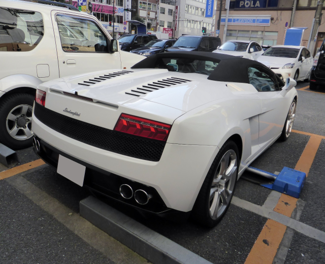 Ein weißer Audi R8 Spyder steht auf einem Parkplatz neben anderen Autos, umgeben von Gebäuden, Schildern, Pfählen, Bäumen und einem klaren blauen Himmel.