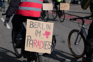 Eine Gruppe von Menschen, die auf Fahrr├Ądern eine Stra├če entlangfahren, mit einem "Berlin I'm Paradies"-Schild im Vordergrund und einem Auto im Hintergrund.
