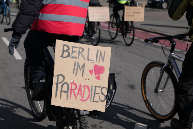 Eine Gruppe von Menschen, die auf Fahrr├Ądern eine Stra├če entlangfahren, mit einem "Berlin I'm Paradies"-Schild im Vordergrund und einem Auto im Hintergrund.