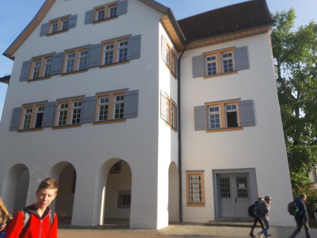 Eine Gruppe von Menschen, einige mit Taschen, geht vor einem großen weißen Gebäude mit Fenstern und Türen vorbei, das von Bäumen umgeben ist und einen klaren blauen Himmel hat.