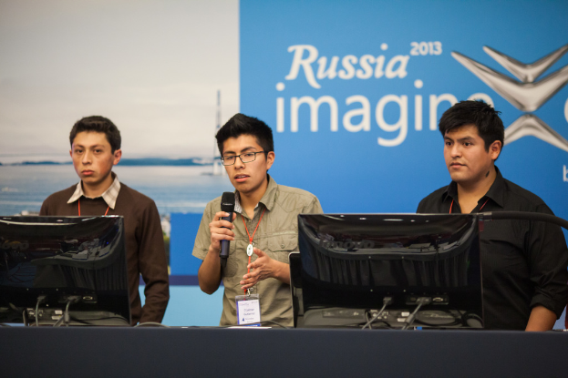 Drei Jungen stehen neben einem Schreibtisch mit zwei Computersystemen, einer spricht in ein Mikrofon, trägt ID-Karten mit roten Tags, vor einer Wand mit einer Werbetafel, auf der "Russia Imagine 2013" steht.