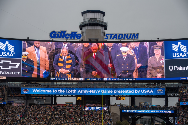 A large crowd in a stadium watches a football game, with a central screen displaying images and text, boards with text, spectators in the stands, an arch, trees, and a clear blue sky in the background.