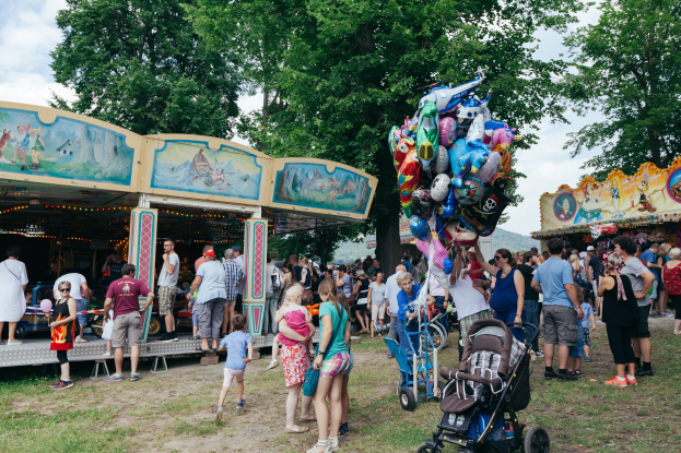 Eine Menschenmenge umringt einen bunten, beleuchteten Karussellwagen auf einem Volksfest, einige halten Kinderwagen, mit Bäumen und einem bewölktem Himmel im Hintergrund.