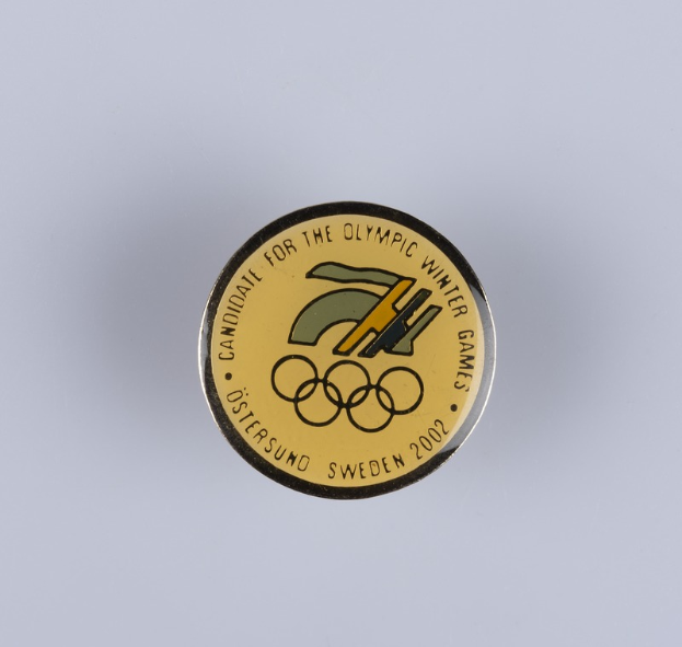Ein rundes Abzeichen mit schwarzem Hintergrund und goldfarbenem Olympische Winterspiele-Logo, mit der Aufschrift "Olympische Winterspiele" in fetter weißer Schrift.