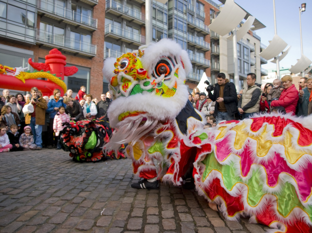 Ein lebendiges chinesisches Neujahrsfest in Amsterdam mit einer Löwendance-Aufführung und einer Menge Schaulustiger, einige halten Kameras, vor einem Hintergrund aus Gebäuden, Laternenmästen und einem klaren blauen Himmel.