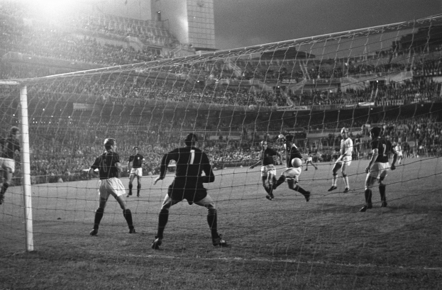 Eine Gruppe von Männern, die Fußball in einem Stadion spielt, mit einem Torpfosten im Vordergrund und einem Gebäude im Hintergrund, dargestellt in Schwarz-Weiß.