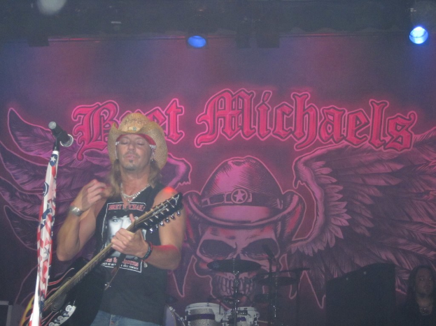 Eine Musik-Konzert-Szene mit Scheinwerfern oben, einem Banner im Hintergrund, Drogen in der Mitte unten, einer Person in der rechten unteren Ecke sitzend und einem Gitarristen mit Mikrofon links.