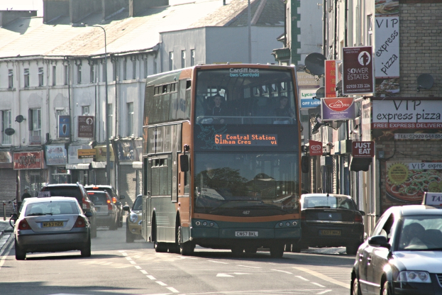 Eine Straße mit Autos und einem Bus vor Gebäuden mit Wänden, Fenstern, Tellern und Dächern, die Plakate und Banner an den Wänden und einen Laternenmast zeigen.