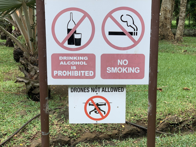 Schankverbot an der Straßenseite mit Text "Alkoholgenuss ist untersagt" und "Rauchen verboten", umgeben von Bäumen und Gras.