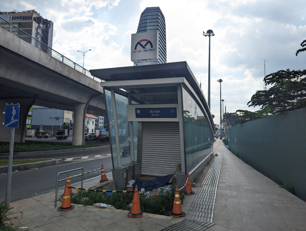 Busbahnhof mit Verkehrskegeln, Pflanzen und einem Schild neben einem hohen Gebäude, Fahrzeugen auf der Straße und einem bewölkten Himmel.