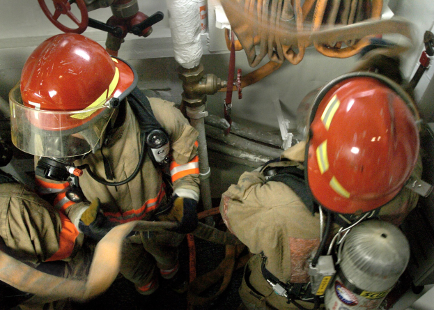 Feuerwehrleute in Schutzausrüstung an einem Hydranten mit Rohren und Ventilen im Hintergrund.