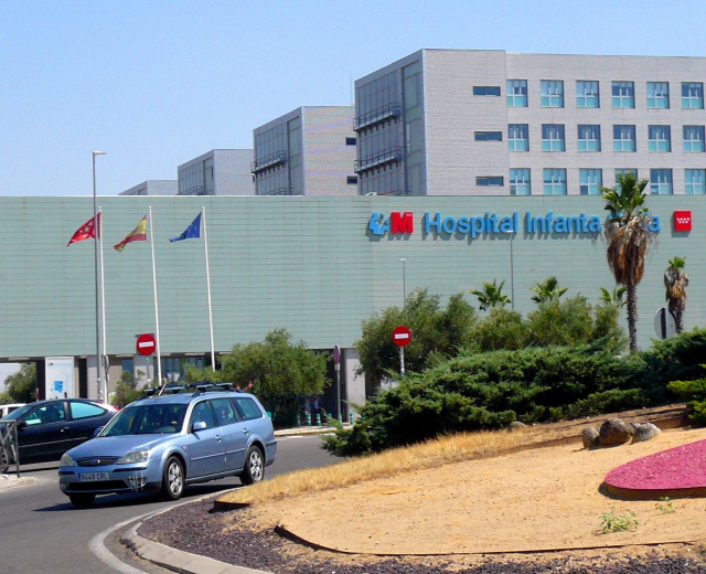 Krankenhaus mit parkenden Autos, umgeben von städtischer Infrastruktur und Grün unter einem klaren Himmel.