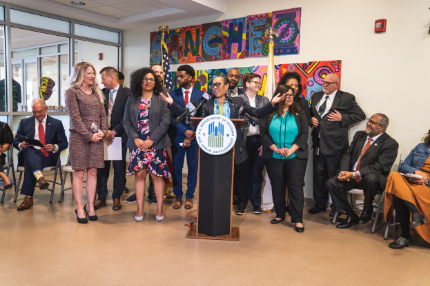Gruppe von Menschen vor einem Rednerpult mit Mikrofon und einer Tafel mit der Aufschrift "Sancho Stadtrat tritt dem Stadtrat bei", mit sitzenden und stehenden Teilnehmern, Flaggen, Feuerlöscher und Deckenleuchten im Hintergrund.