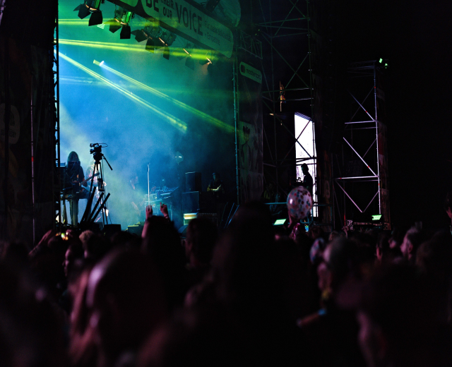 Ein volles Konzert mit Musikern auf der Bühne, beleuchtet von Scheinwerfern, vor einem dunklen Hintergrund.