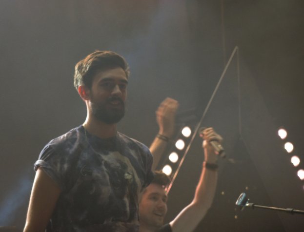 Ein Mann in einem T-Shirt steht und lacht, mit einem anderen lachenden Mann, Lichtern, Rauch, einem Mikrofon und anderer Ausrüstung im Hintergrund.