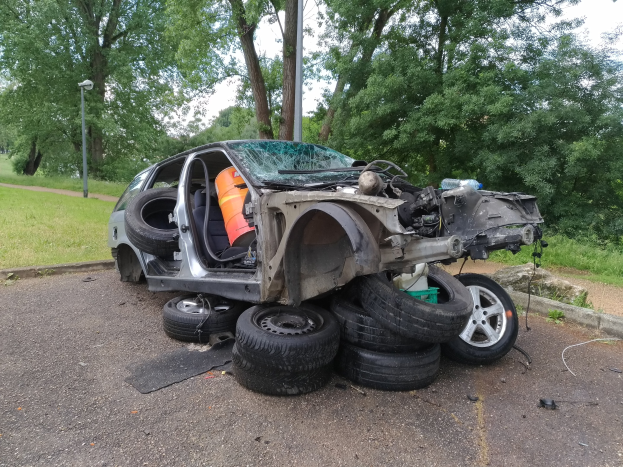 Ein umgestörter Wagen am Straßenrand mit verstreuter Trömmern, umgeben von Gras, Pflanzen, Bäumen, einem Laternenpfahl und einem bewölktem Himmel.