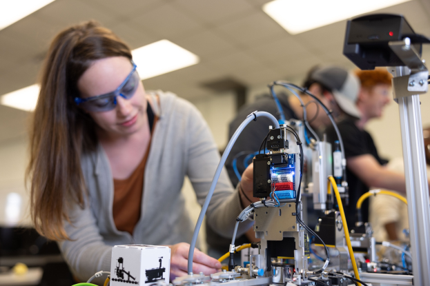 Eine Frau in Schutzbrille arbeitet an einem Roboter in einem Labor umgeben von anderen, mit Gegenständen auf dem Tisch und Deckenbeleuchtung.