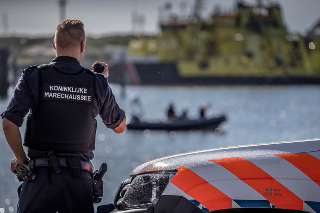 Ein Polizeibeamter in Uniform steht neben einem Polizeiwagen in der Nähe eines Gewässers und hält ein Funkgerät in der Hand, im Hintergrund ist ein Boot mit ein paar Menschen auf dem Wasser zu sehen und der Himmel darüber.