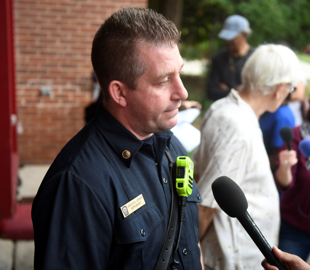 Ein Polizist in Uniform spricht zu Reportern im Freien und hält ein Mikrofon, während andere mit Mikrofonen in der Nähe stehen.
