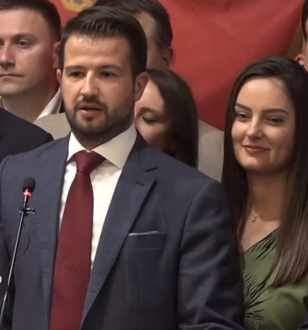 Ein Mann im Anzug spricht in ein Mikrofon an einem Podium, mit einem Banner dahinter, zu einer Menge.