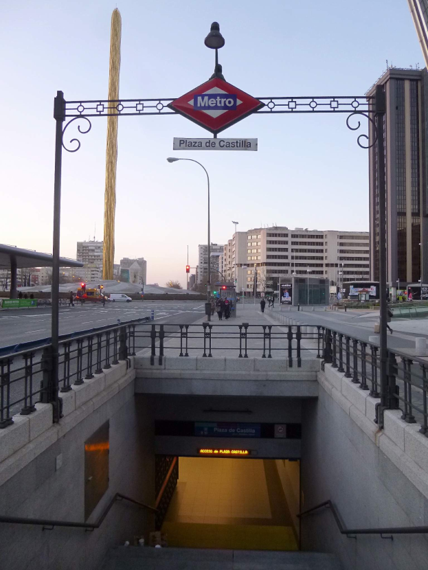 Eine belebte Stadtkulisse mit Gebäuden, Fahrzeugen, Passanten, einem U-Bahn-Eingang und einer darüber befindlichen Tafel.