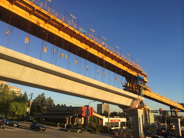 Eine Baustelle in der Mitte mit Fahrzeugen und Gebäuden auf der linken Seite, unter einem blauen Himmel.