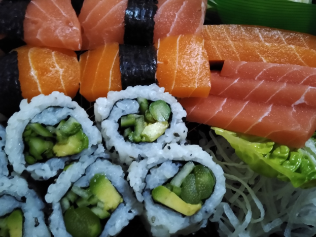 Ein schwarzer Teller mit Lachs, Avocado und Gurken-Sushi, umgeben von ein paar grünen Blättern.