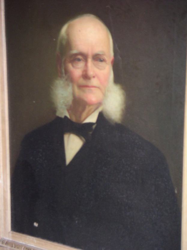 Ein Schwarz-Weiß-Porträt eines älteren Mannes mit einem weißen Bart und Brille, der schlichte Kleidung trägt und in einem Foto Rahmen steht.