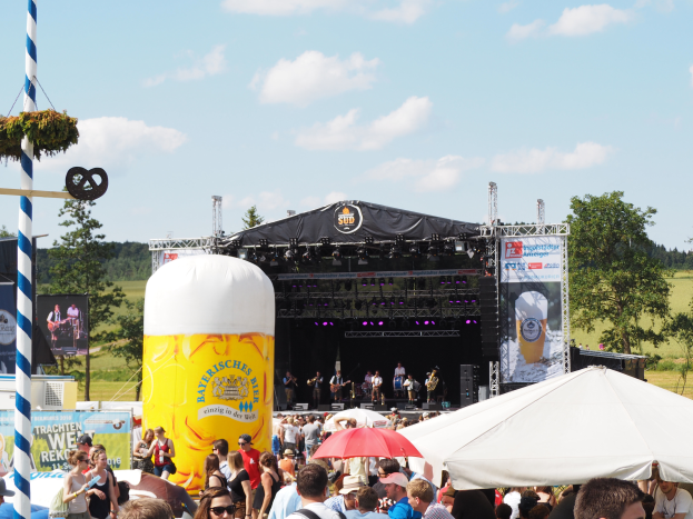 Eine große Menschenmenge beim Oktoberfest in München, viele mit Mützen und Goggles, einige mit Schirmen, ein Zelt im Vordergrund, ein dekorierter Pfahl links, Bäume und ein bewölkter Himmel im Hintergrund und eine Bühne mit einem Banner und Musikern.