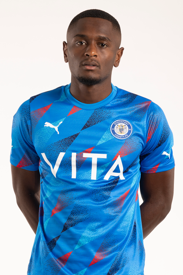 Ein Mann in einem blauen Leicester City Away-Shirt von der Saison 2018-19, der entspannt mit Händen in den Taschen und einem leichten Lächeln posiert.