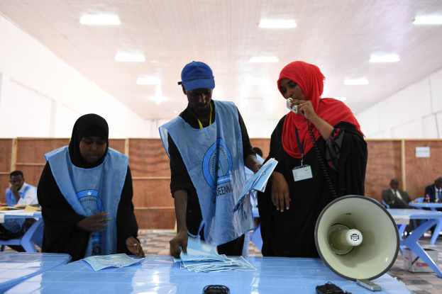 Menschen versammeln sich um einen Tisch mit Megafon und Papieren an einer Wahllokal, während andere im Hintergrund auf Stühlen sitzen und stehen.