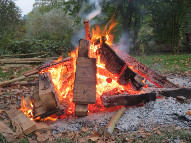 Lagerfeuer in einem bewaldeten Gebiet umgeben von Holzscheiten, Steinen und getrockneten Blättern mit Bäumen und einem klaren blauen Himmel im Hintergrund.