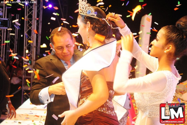 Ein Mädchen auf der Bühne bei einer Miss-World-Show, das eine Krone trägt, während ein anderer Mädchen die Krone hinter ihr anpasst und ein Mann in einem schwarzen Anzug vor ihr die Hand schüttelt, vor einem Hintergrund aus Metallrahmen und einer dunklen Kulisse.