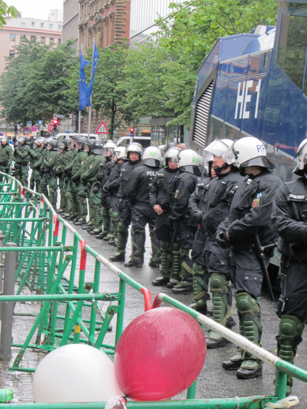 Eine Gruppe von Polizisten in Helmen und mit Waffen steht vor einer Absperrung auf einer Straße mit Geländern und Luftballons, mit Bäumen, Gebäuden, Flaggen und einem klaren blauen Himmel im Hintergrund.