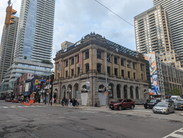 Eine belebte Stadtstraße mit hohen Gebäuden im Hintergrund, Fahrzeugen, Fußgängern, Verkehrsinfrastruktur und Bäumen, zentriert auf der Toronto Public Library.