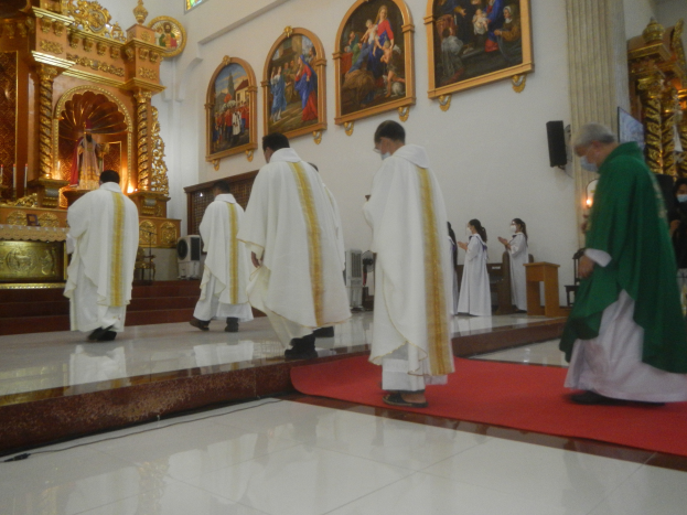 Eine Gruppe von Priestern geht einen roten Teppich in einer Kirche entlang, während Menschen auf dem Boden vor und rechts von ihnen stehen und Fotorahmen an der Hintergrundwand hängen.