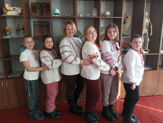 Eine Gruppe von Kindern in traditioneller ukrainischer Kleidung steht vor einem Regal, das mit Spielzeug, einem Skelett und anderen Gegenständen gefüllt ist, was auf eine ukrainische Schulumgebung hinweist.