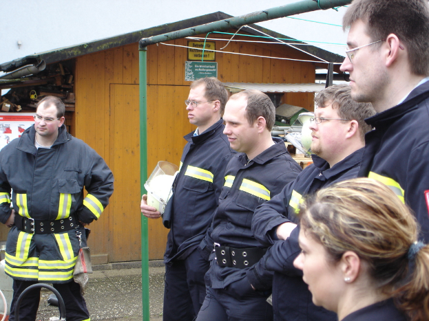 Eine Gruppe von Männern in blauen Feuerrettungsuniformen, die nach links schauen und vor einem kleinen Holzschuppen und Rohrrahmen stehen.