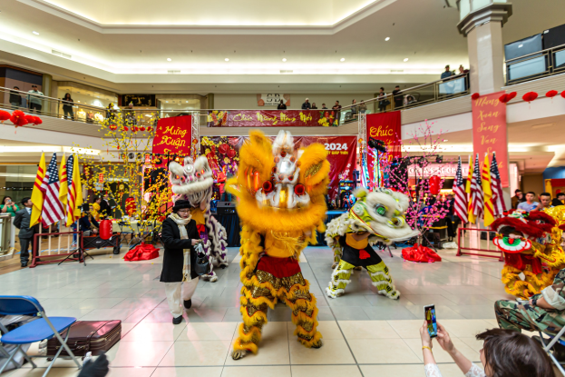Ein lebendiges chinesisches Neujahrsfest in einem Einkaufszentrum mit Menschen in Kostümen, die tanzen, andere auf Stühlen sitzen und Handys halten, sowie Flaggen, Bannern, Dekorationen und Deckenleuchten im Hintergrund.