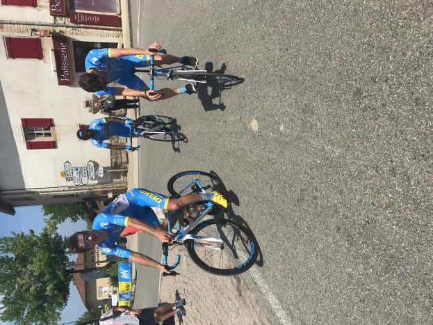 Eine Gruppe von Radfahrern fährt eine Straße entlang an einem Gebäude mit Fenstern und Schildern vorbei, mit Bäumen und einem blauen Himmel im Hintergrund und Zuschauern, die auf der Straße darunter stehen.