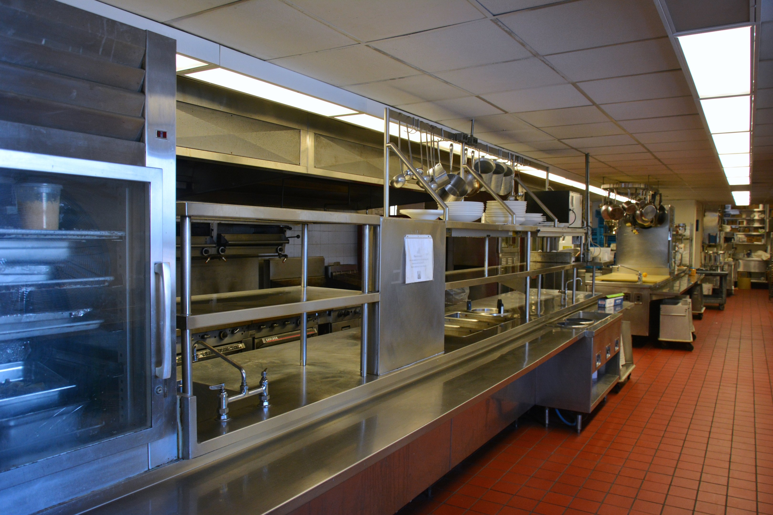 Kommerzielle Küche mit Edelstahl-Theke, hängenden Utensilien, einem Spülbecken, Schränken und verschiedenen auf Gestellen angeordneten Gegenständen unter elektrischem Licht.