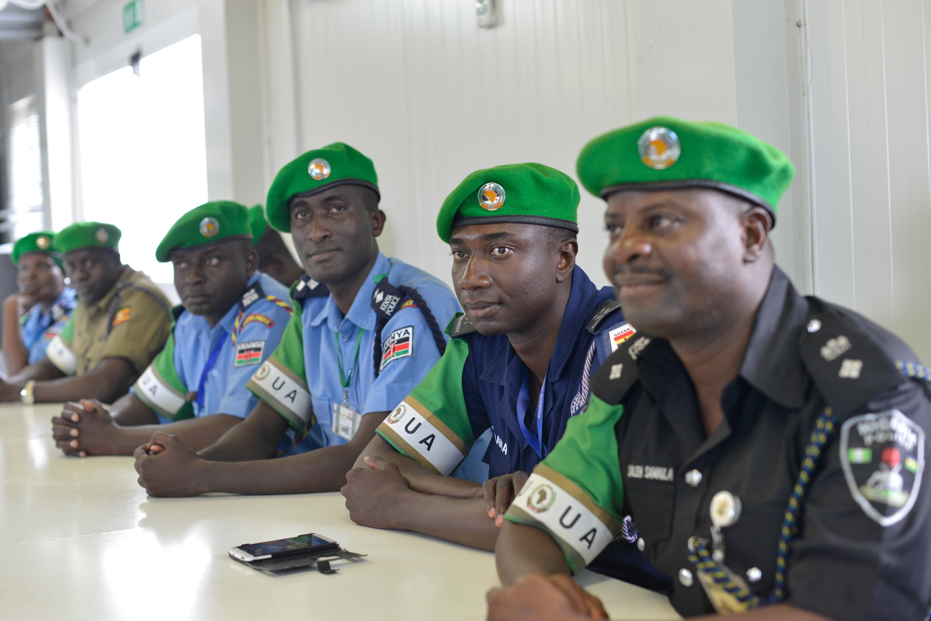 Gruppe von Polizeibeamten, die an einem Tisch während einer Schulung sitzen, mit Mobiltelefonen und anderen Gegenständen auf dem Tisch, in einem Raum mit Fenstern und Deckenbeleuchtung.