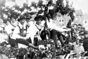 Eine Gruppe von Menschen in traditioneller bayrischer Tracht fährt auf einem blattgeschmückten Festwagen in einem Umzug mit, einige halten Musikinstrumente, mit Gebäuden und einem klaren Himmel im Hintergrund, dargestellt in Schwarz-Weiß.